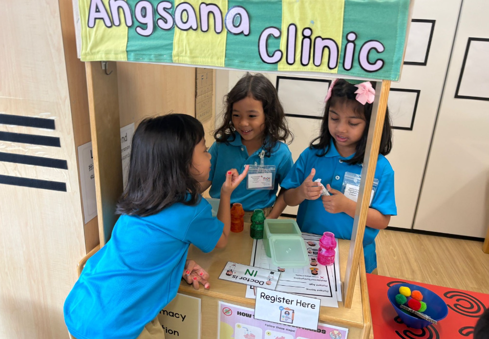 Three children in blue shirts play at a booth labeled Angsana Clinic, with a Register Here sign.